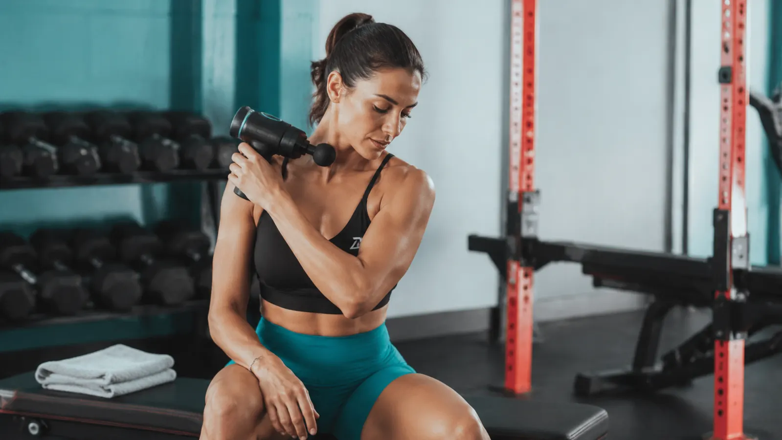 Woman using a percussion massage gun on her upper back after a workout, gym bag nearby