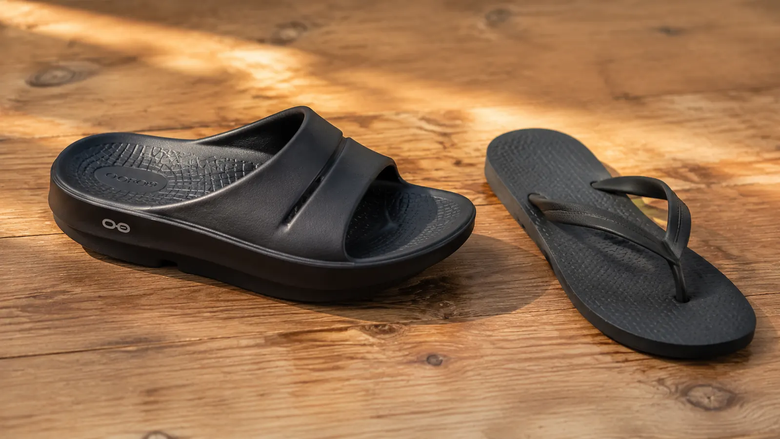 Side-by-side comparison of an OOFOS OOahh slide and a standard foam flip-flop on a white surface showing the dramatic thickness and arch contour difference