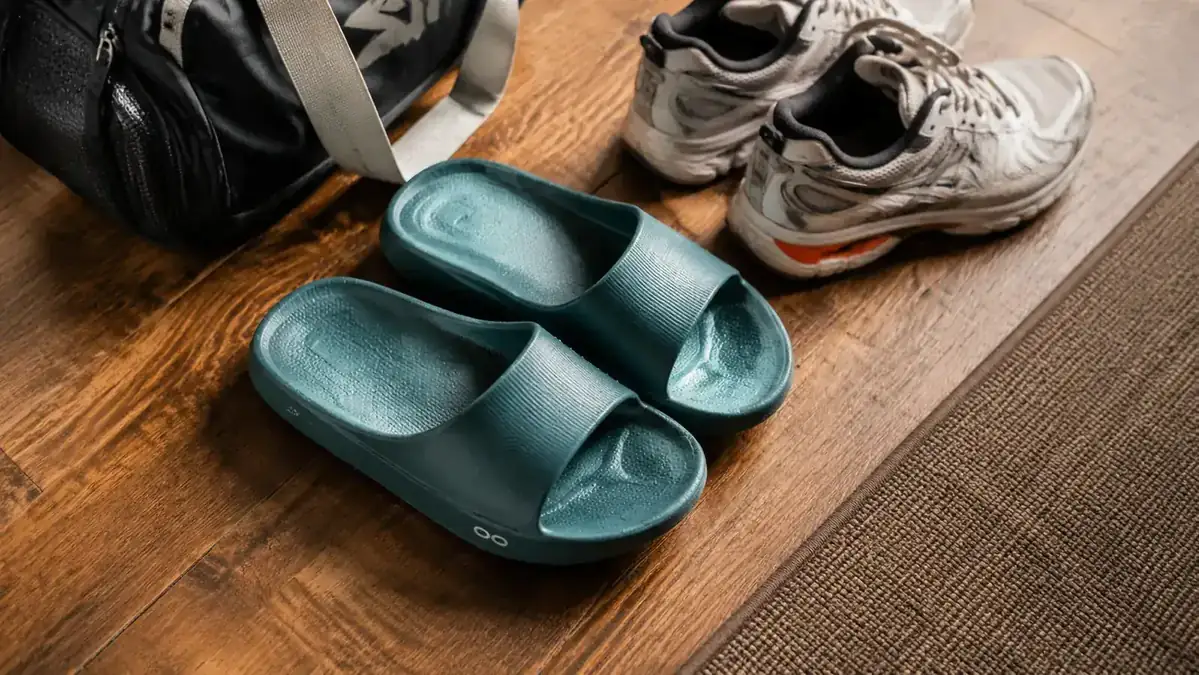 A pair of OOFOS OOahh recovery slides resting on a hardwood floor next to a pair of running shoes and a small gym bag