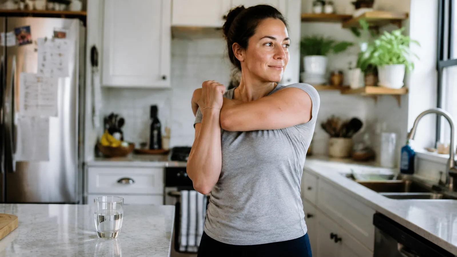 Person doing a light morning stretch in a bright kitchen while holding a glass of water after taking a supplement